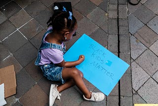 A young Black girl sits on the ground with a bright blue sign that says “Black Kids Matter.”