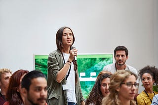 A woman speaks into a microphone, asking questions while standing amidst colleagues.