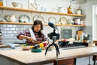 Adult woman preparing a meal and chopping fresh ingredients in front of a video camera on a tripod.