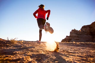 A photo of a woman running on a desert trail, her left foot kicking up dirt.