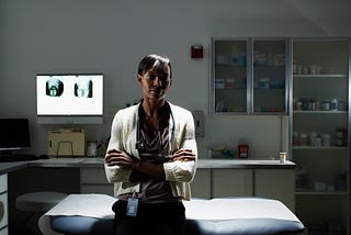 A photo of a Black female doctor sitting in an examination room, arms crossed.
