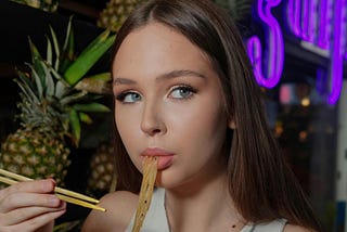 A young woman eating vegetable noodles with pineapples in the background