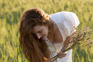 Red-haried woman with long hair bends down to gather a bunch of long blooming grass on a very sunny day. She is dressed in an airy white dress. You can see down her blouse a bit, but the picture is unprovocative. You see long grass all around here.