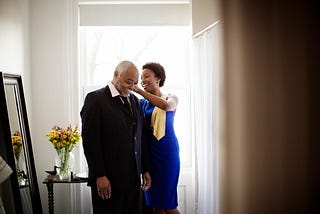 A Black woman assists her elder father with his collar as they prepare for a party.