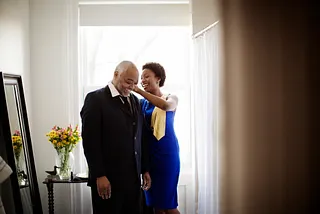 A Black woman assists her elder father with his collar as they prepare for a party.