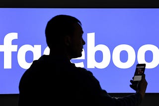 A man with a smartphone stands in front of a monitor with the Facebook logo.