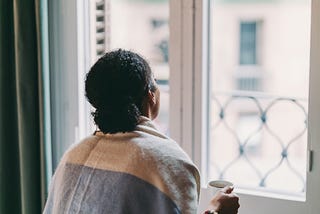 Black woman holding a cup of coffee while looking out her window.