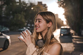 A smiling woman is standing on a street while holding a cup of coffee.