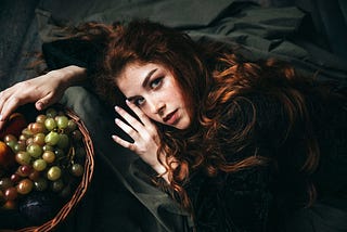 A woman wearing a vintage dress is lying on the floor with a basket of grapes.