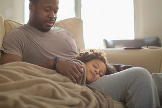 A photo of a young black girl sleeping with her head on her dad’s lap.