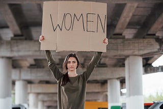 A woman stands beneath an overpass. With her hands, she’s holding high a cardboard sign with the word WOMEN on it.