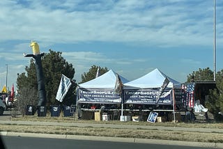 Two Trump booths are side by side next to a road. There are American flags hanging from them beside signs that say “TrumpShopUS.com: Keep America Great”.