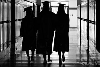 A black and white picture of three high school grads walking down the school hallway wearing their grad gowns.