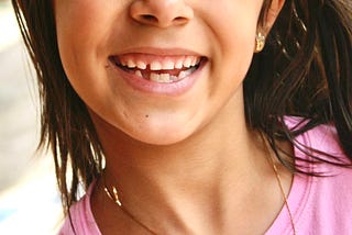 Color square closeup photo of a tanned Costa Rican girl’s smiling face, where we see the bottom half with her nose and camera-centered on her big grin, with the lower layer of her teeth semi-toothless, dark black straight shoulder-length hair in the background, and seeing specks of a cotton-candy pink t-shirt and thin gold necklace with a profiled-praying-to-the-left shiva charm.