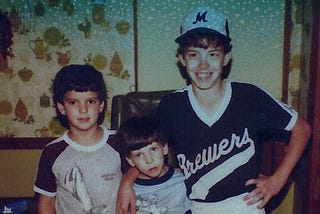 Three young boys pose for a Polaroid picture standing against a wall with 1970s-era wallpaper.