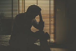 A silhouette of a man sitting on his bed with his hand against his forehead, seemingly upset, light seeping from the blinds.