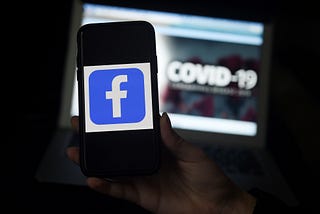 Facebook logo is displayed on a mobile phone screen with the words COVID-19 blurred in the background on a laptop screen