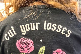 Color photo showing the back of the hair stylist’s black t-shirt, upon which is the wording “cut your losses” with a graphic image of a skeleton hand holding a pair of scissors that are snipping 2 pink roses from their stems.