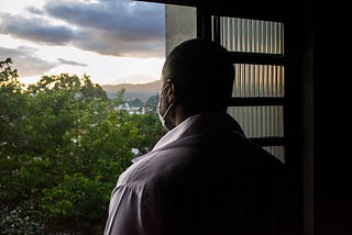 A photo from the back of a black man wearing a face mask looking out his window.