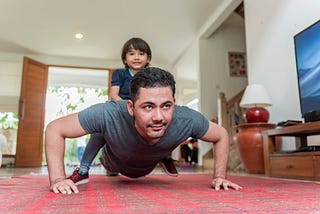 A photo of a father doing push ups in the living room while his son sits on his back.