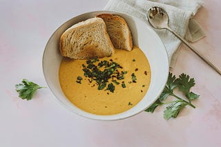 A bowl of creamy yellow soup garnished with chopped fresh parsley sits on a light-colored surface. Two toasted white bread rest on the edge of the white bowl, and a silver spoon lies nearby on a linen napkin.