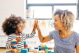 A photo of a mom and her young child high fiving as the kid plays with blocks.
