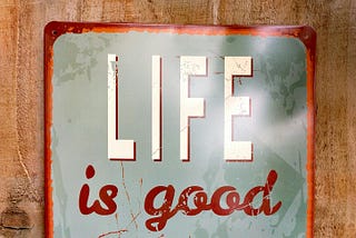 Vintage-style decorative sign mounted on a wooden surface. The sign has a weathered light blue-green background with rust-colored edges. The text reads “Life is good at the beach”. The design evokes a relaxed, coastal vibe, ideal for beachside decor or vacation homes.