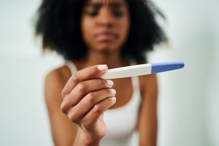 Black woman holding a pregnancy test with an upset expression.