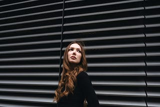 A woman wearing a chic black outfit is leaning against a gray, shuttered wall.