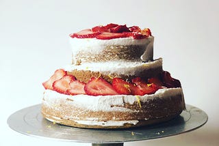 A layered cake on a silver cake stand — the cake is golden brown with lightly applied white icing and strawberries.