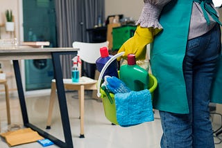 A waist-down shot of a maid, seen from behind, holding a bucket of cleaning products in a small room.