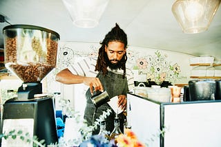 A Black barista, their long locs tied back, pours milk into a coffee cup behind the counter of a coffee truck.