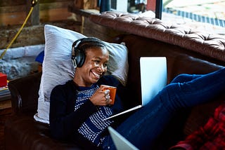 Smiling Black woman with headphones on looking at her computer with a cup of coffee in her hand.