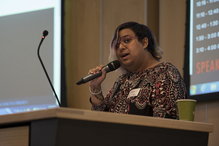 A woman speaking into a mic, standing at a podium wearing a patterned dress.