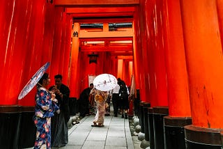 The overwhelmingly red view of Fushimi Inari Shrine in Kyoto, Japan. Two woman are dressed in traditional kimonos with outfit matching umbrellas. A man, also in traditional garb, is taking a photo of one of the women.