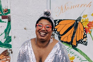 Sondra — a Black woman — poses in a sparkling silver gown. Behind her is a mural showing butterflies in bright colors.