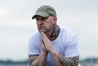 Man sitting outside on a rock looking lost in thought and a bit sad. Pensive. He is wearing a white t-shirt and jeans, with a green ballcap. His arms are tattooed. He has workboots on. The background is blurred but looks like a beach’s rocky shore.