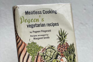 A hand holding up a vintage cookbook with a quaint illustration of fruits and veggies on the front.