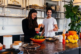 Two smiling young adults in a fancy kitchen. One, probably a woman, is grinding pepper onto a salad.