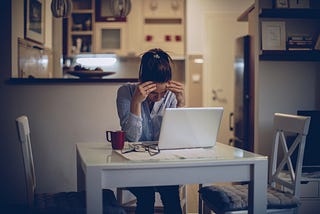 One woman with her head in her hands, sitting alone at home, working on laptop.