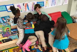 A smiling teacher sitting on a small couch under a pile of students who are hugging them.