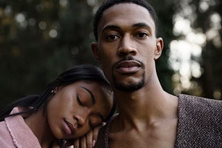 A photo of a black woman resting her head on the shoulder of a black man who is looking at the camera.