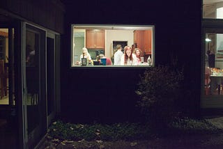 Family group washing dishes in the kitchen after a Thanksgiving meal, shot from outdoors through a picture window at night.