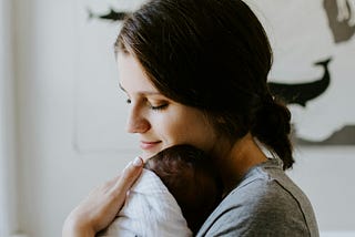 A dark haired woman holding a sleeping little baby in her arms. her chin is resting on his forehead .
