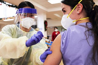 A healthcare worker wearing a face mask and a face shield giving an injection to another masked healthcare worker.