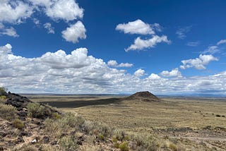 A mostly flat New Mexican landscape with scrubby grass under a deep blue sky with white fluffy clouds.