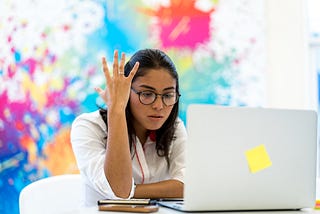 A woman throws her arm up in frustration as she stares at her laptop.