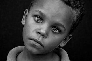 A black and white photo of a young, dark-skinned boy with his head tilted to one side and looking directly at the camera. The words, ‘the miracle of sight’ are repeated 4 times under the image in font colours that graduate from white to dark grey.