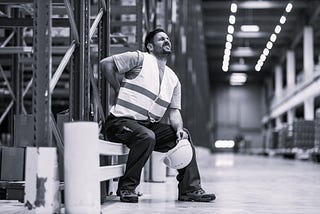Man sitting in warehouse with a painful grimace on his face while holding his back. He is wearing a work vest and holding a hard hat.