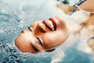“Embodiment and Reconnecting to Self: Embrace Inner Harmony” This photo shows the face of a woman with carmel-brown skin floating in a pool of blue water. Only her face is above the water and she is smiling.